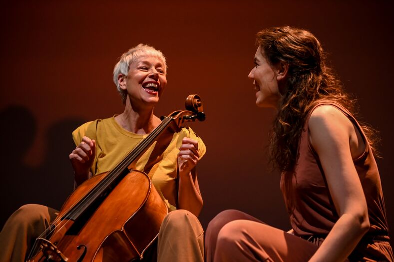 Twee vrouwen lachen en zitten samen op een podium, één met een cello en de ander in een bruine outfit, tegen een warme achtergrond.