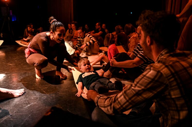 Groep mensen op een podium tijdens een voorstelling met een baby in het midden, terwijl volwassenen interactie hebben.