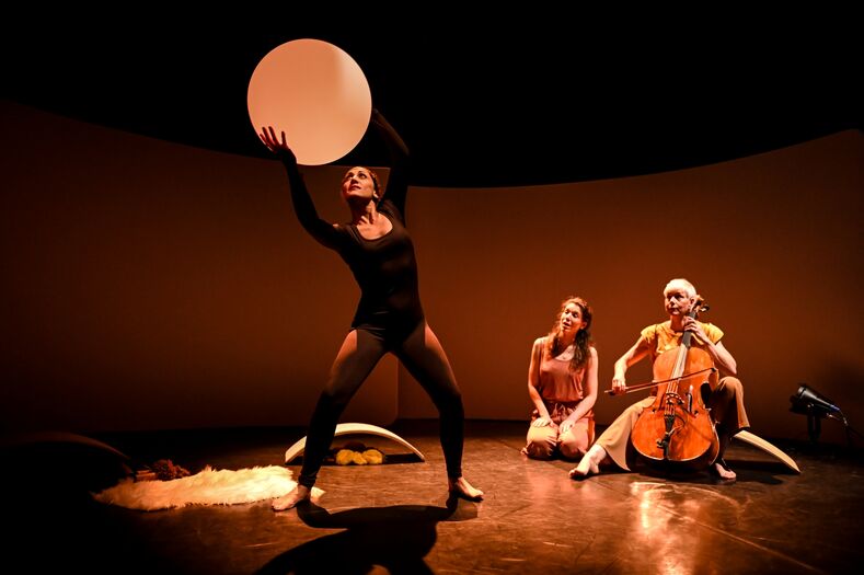Een performer in zwart kostuum houdt een ronde schijf omhoog, terwijl twee andere artiesten toekijken, eentje met een cello.