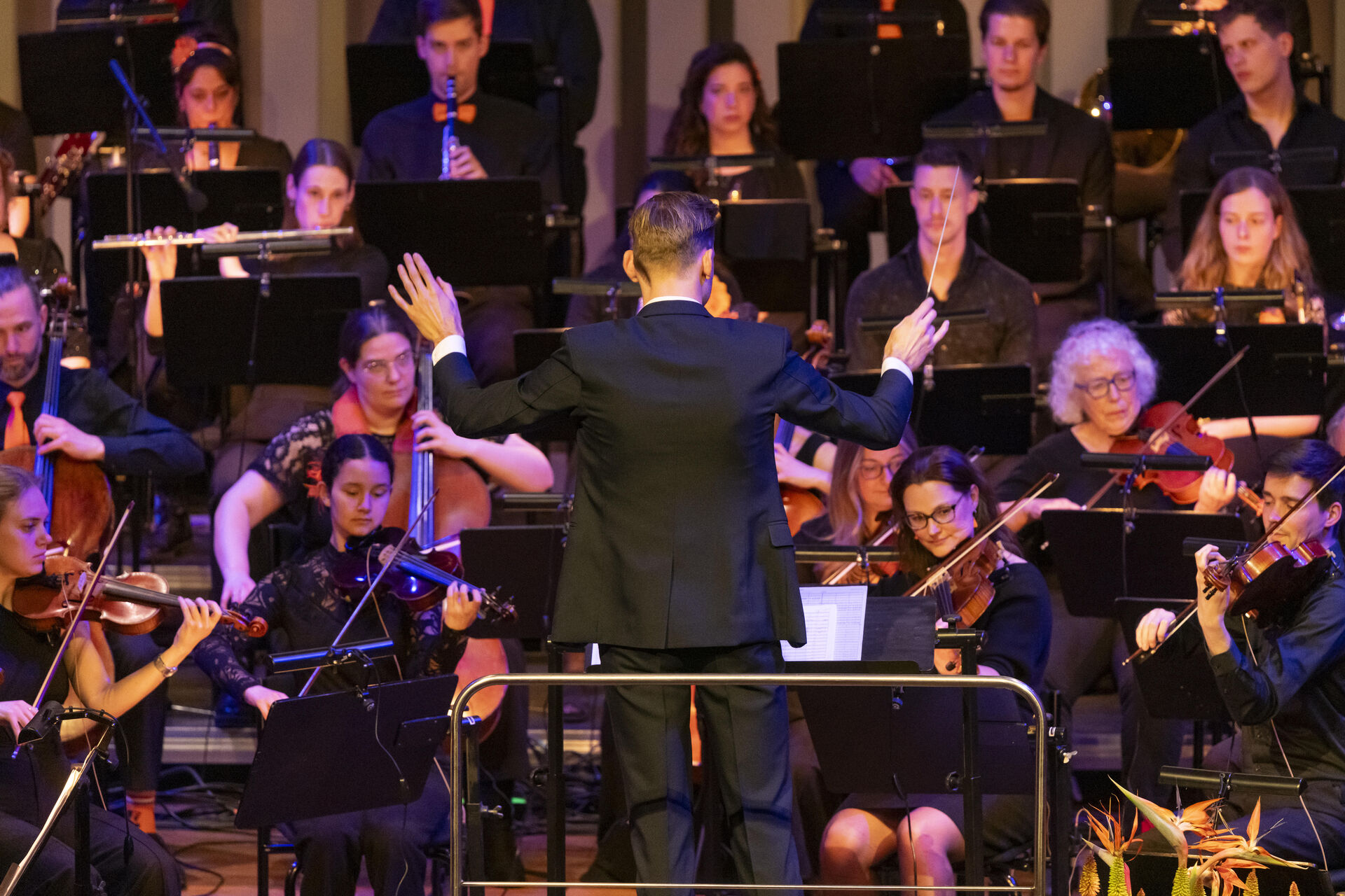 Een dirigent leidt een orkest tijdens een optreden, met muzikanten die verschillende instrumenten bespelen.
