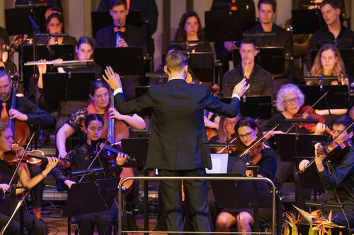 Een dirigent leidt een orkest tijdens een optreden, met muzikanten die verschillende instrumenten bespelen.