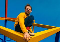 Martijn Crins in gele trui en donkerblauwe broek zit balancerend op een felgele constructie tegen een effen blauwe achtergrond.