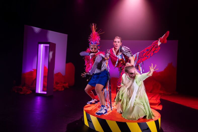 Drie performers in felgekleurde kostuums poseren op een rond platform onder theatraal licht.