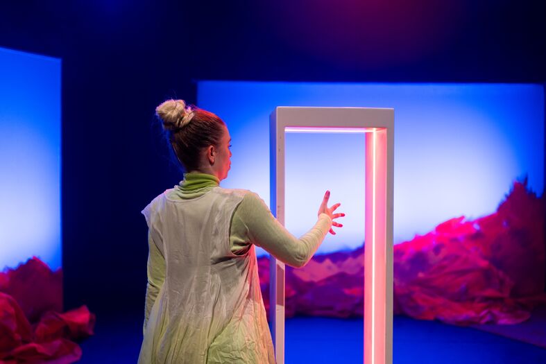 Performer in groene outfit staat voor een deuropening op een podium met felblauw toneellicht en ornaje rotsen.