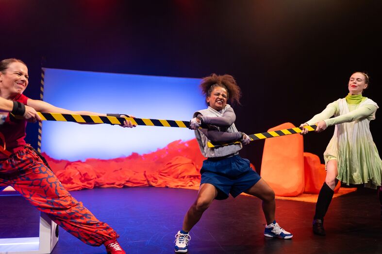Drie performers vechten om een afzetbalk op een podium met felgekleurde oranje objecten.