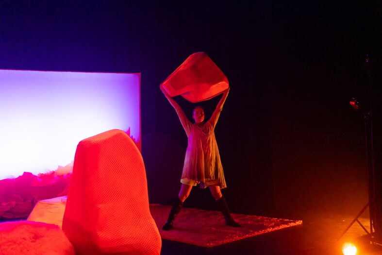 Performer in lichtgekleurde outfit staat op een donker podium en houdt een groot rood object boven het hoofd, omringd door fel oranje decorstukken en paars achterlicht.