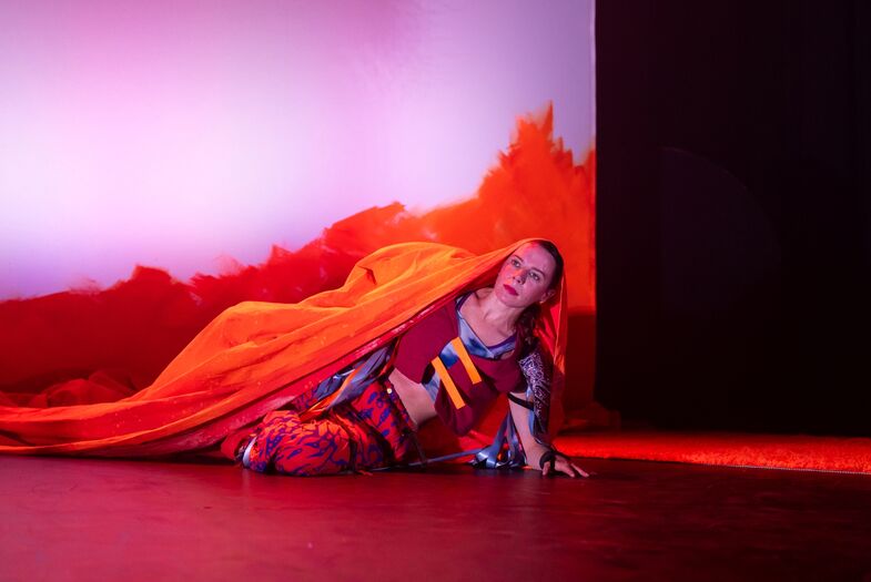 Eén performer ligt op de vloer terwijl een tweede performer het been vasthoudt, met een derde performer rechtop op de achtergrond tussen fel oranje decorstukken