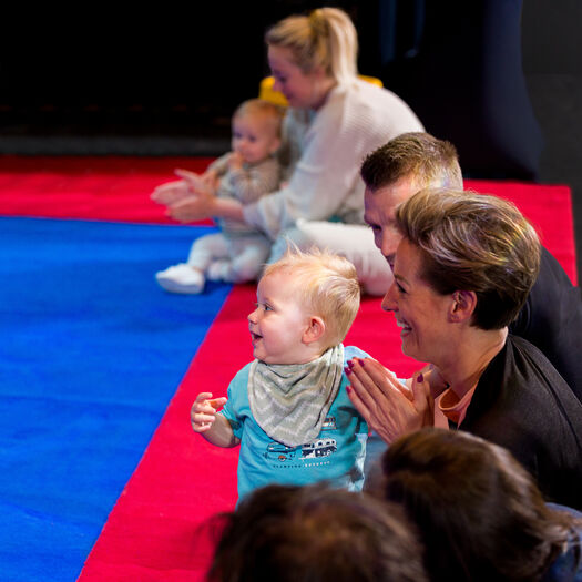 Een kind staat op een mat, omringd door ouders die enthousiast applaudisseren.