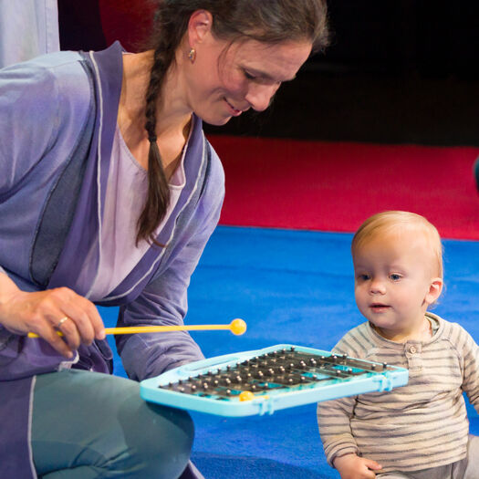 Een vrouw speelt met een baby op een kleed, terwijl ze een muzikale speelgoedtool gebruikt.