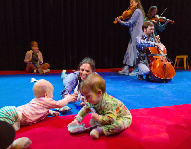 Peuters en ouders spelen op een bontgekleurde vloer terwijl muzikanten op de achtergrond live muziek spelen.
