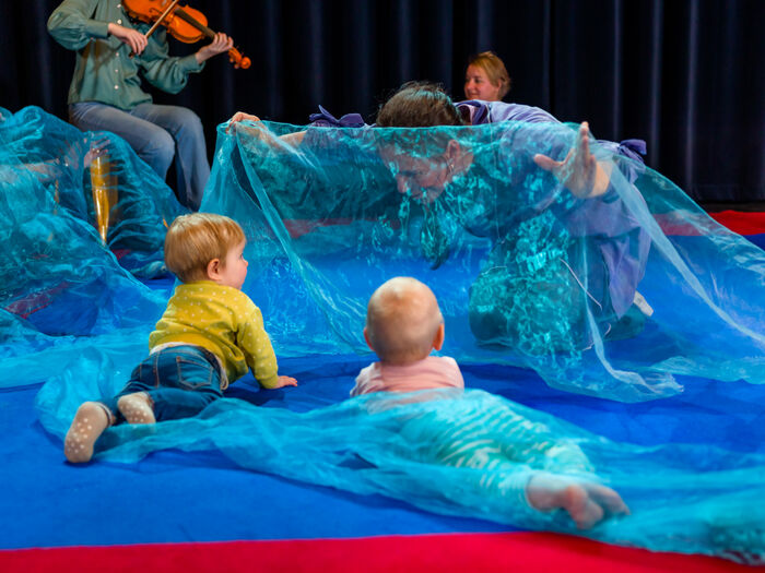 Kinderen spelen op de grond met kleurrijke stof terwijl een violist speelt op de achtergrond.