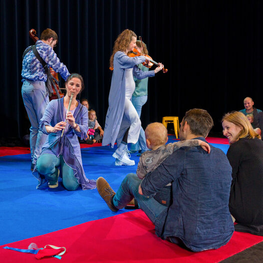 Een groep muzikanten speelt op een podium terwijl kinderen en volwassenen op de grond naar hen kijken.