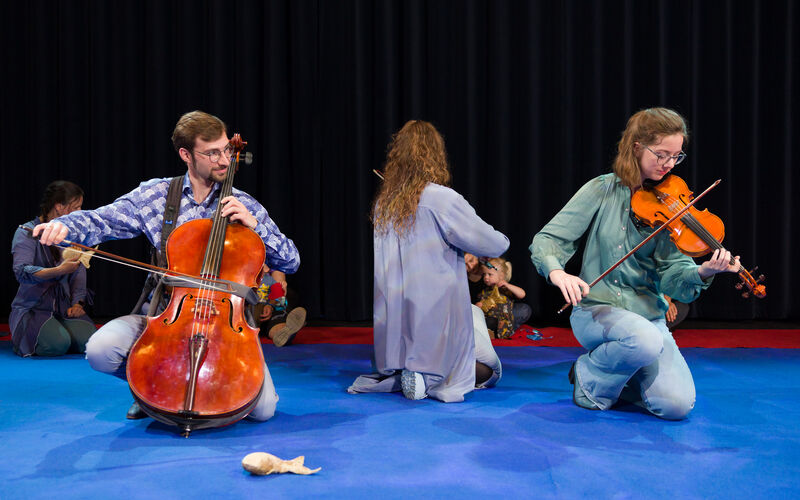 Drie muzikanten in casual kleding spelen samen op een podium, met een cellist, violist en een vrouwelijke violist.