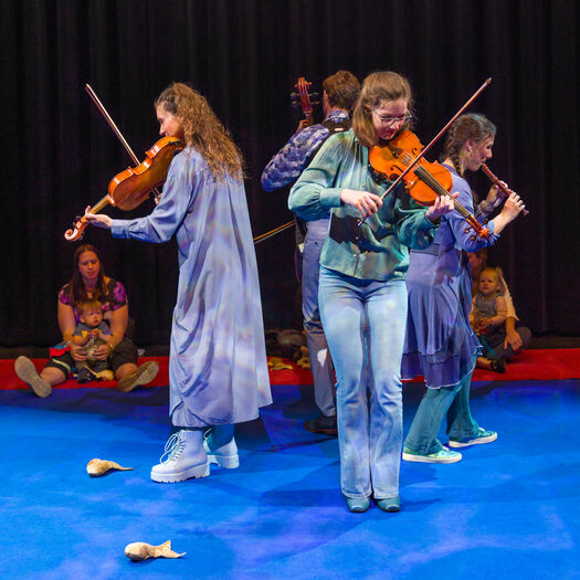 Een groep muzikanten speelt op een podium; twee violisten staan centraal terwijl andere leden achtergrondmuziek bieden.