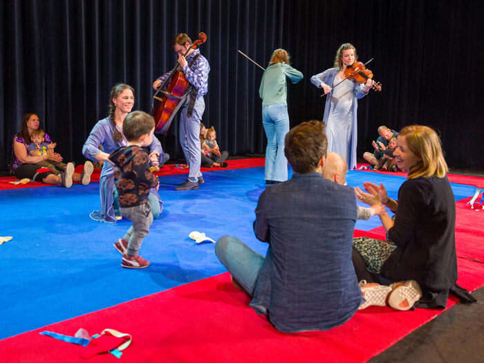 Kinderen en volwassenen genieten van een muziek- en dansvoorstelling, met muzikanten in passende kleding op een kleurige vloer.