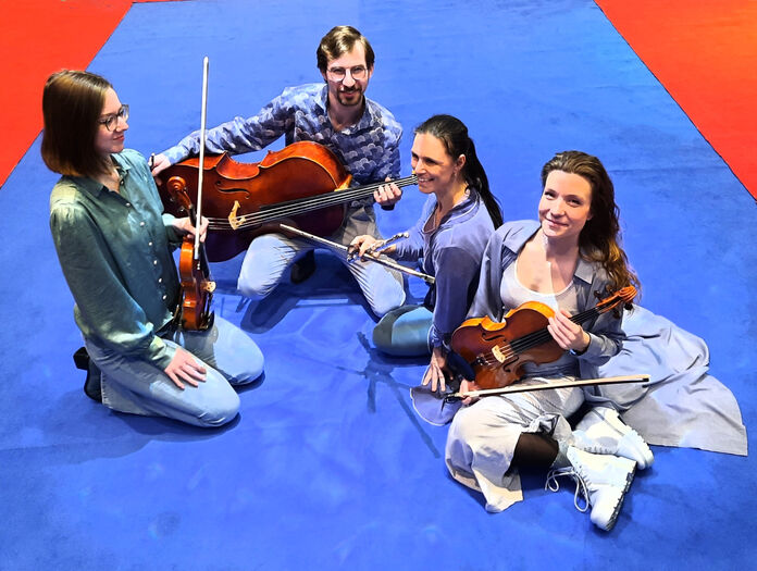 Vijf muzikanten zitten op een blauwe ondergrond met strijkinstrumenten en lachen naar de camera.
