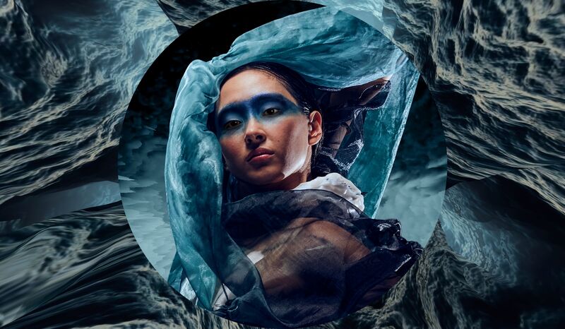 Persoon met blauwe oogmake-up, omhuld door doorschijnende stoffen binnen ronde kadering tegen waterachtig decor.