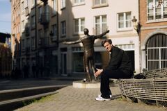 Rob Kemps in donkere kleding zit op een metalen bank in een stadse straat, met een groot bronzen standbeeld met uitgestrekte armen op de achtergrond.