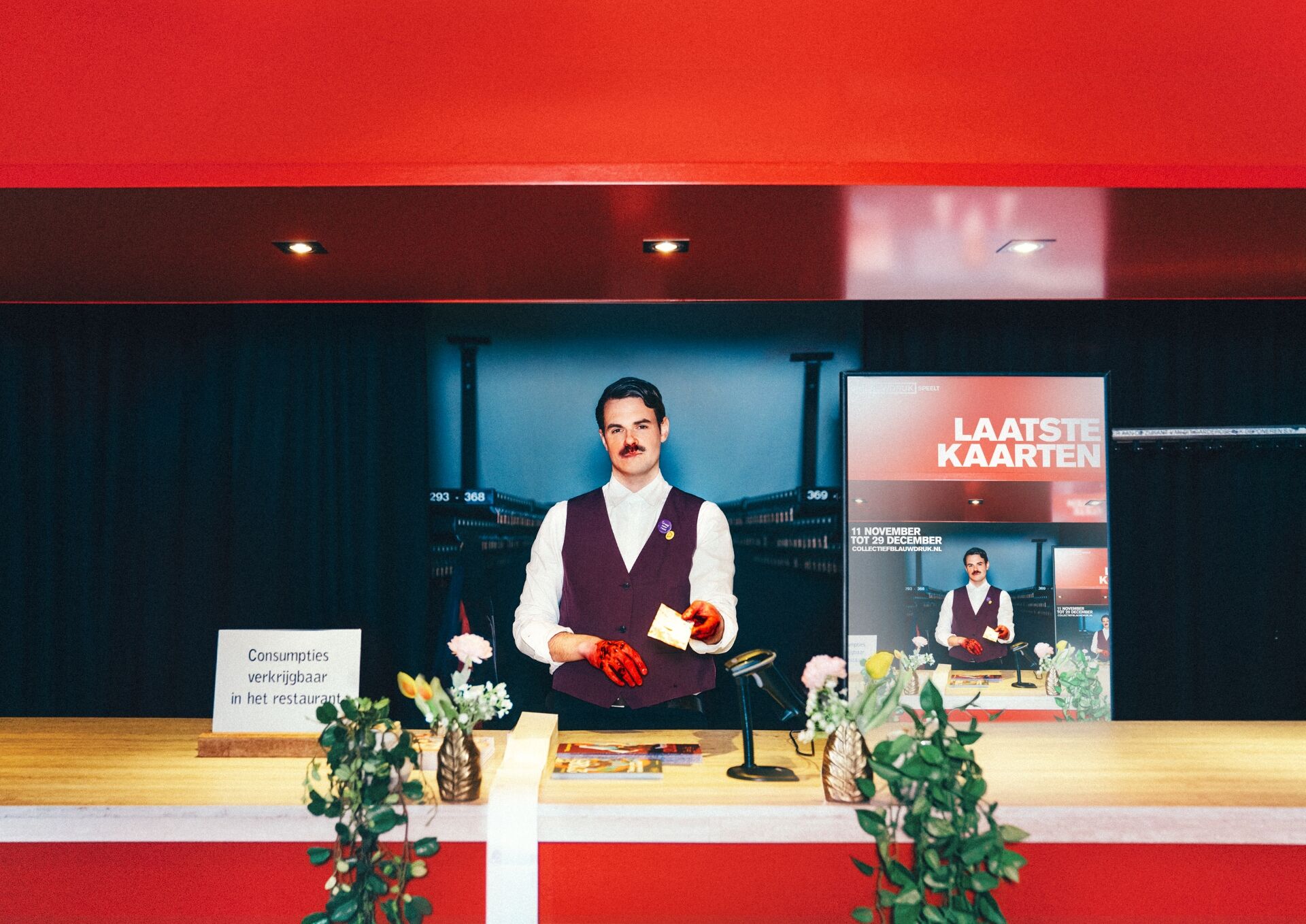 Man in een bordeauxkleurig vest staat achter een balie met planten, terwijl een promotieposter op de achtergrond zichtbaar is.