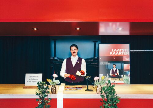 Man in een bordeauxkleurig vest staat achter een balie met planten, terwijl een promotieposter op de achtergrond zichtbaar is.