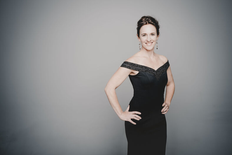 Een vrouw in een elegante zwarte jurk staat met handen op de heupen en glimlacht, tegen een lichte achtergrond.