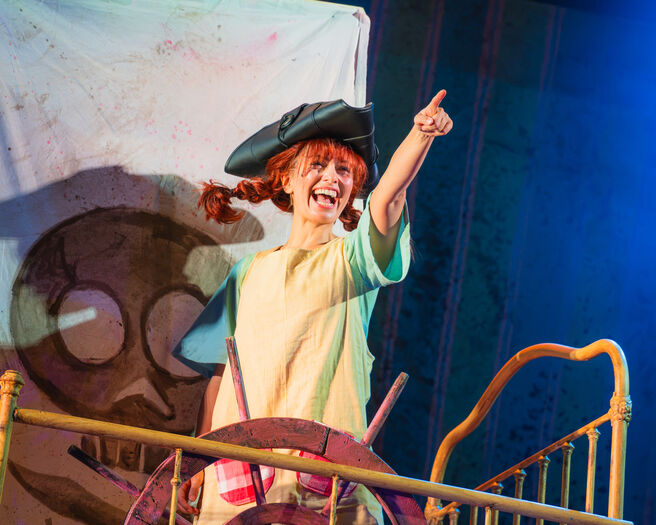 Actrice in een piratenoutfit met een hoed, die enthousiast wijst terwijl ze op een schip staat in een theaterproductie.