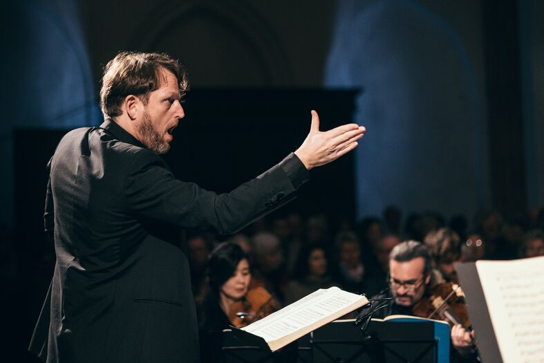 Man in zwart kostuum dirigeert een orkest, met publiek op de achtergrond, tijdens een muziekoptreden.