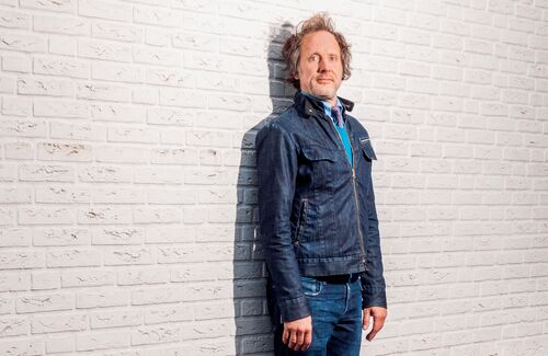 Man in een spijkerjack staat tegen een witte bakstenen muur, met een neutrale houding en lichte glimlach.