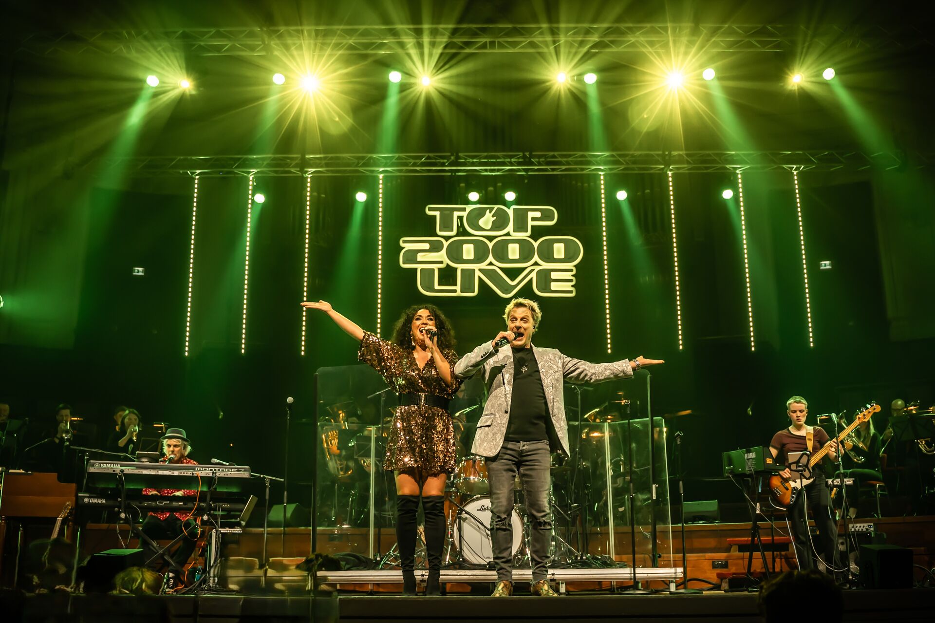 Zanger en zangeres op het podium met band, omringd door groene lichten tijdens een concert.