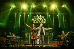 Zanger en zangeres op het podium met band, omringd door groene lichten tijdens een concert.