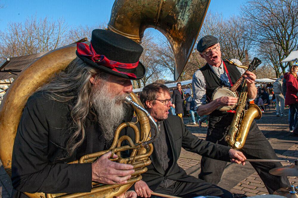 Drie muzikanten in historische kleding spelen samen op straat, met een tuba, een banjo en een saxophone.