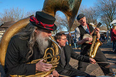 Drie muzikanten in historische kleding spelen samen op straat, met een tuba, een banjo en een saxophone.