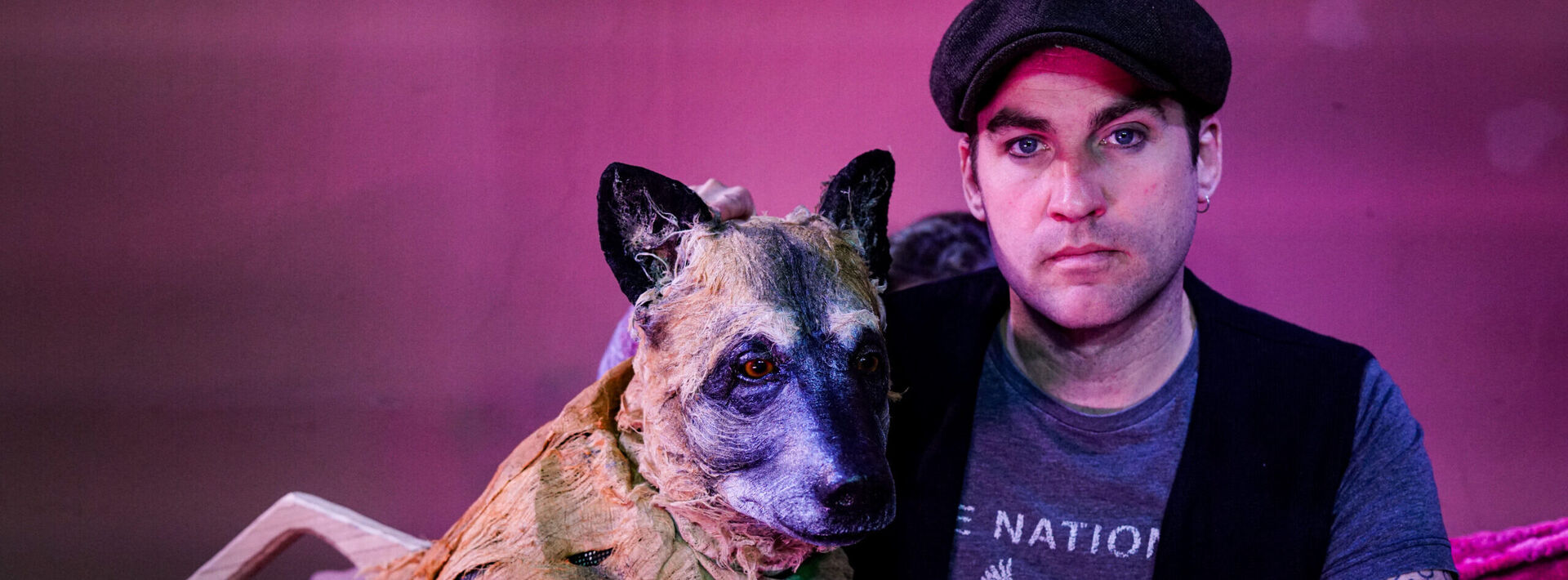 Man met een pet naast een hond met een kostuum, beide poseren voor een paarse achtergrond.