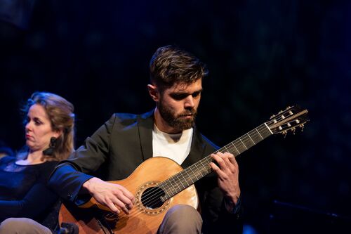 Man met een gitaar speelt op een podium, terwijl een vrouw naast hem op de achtergrond zit.