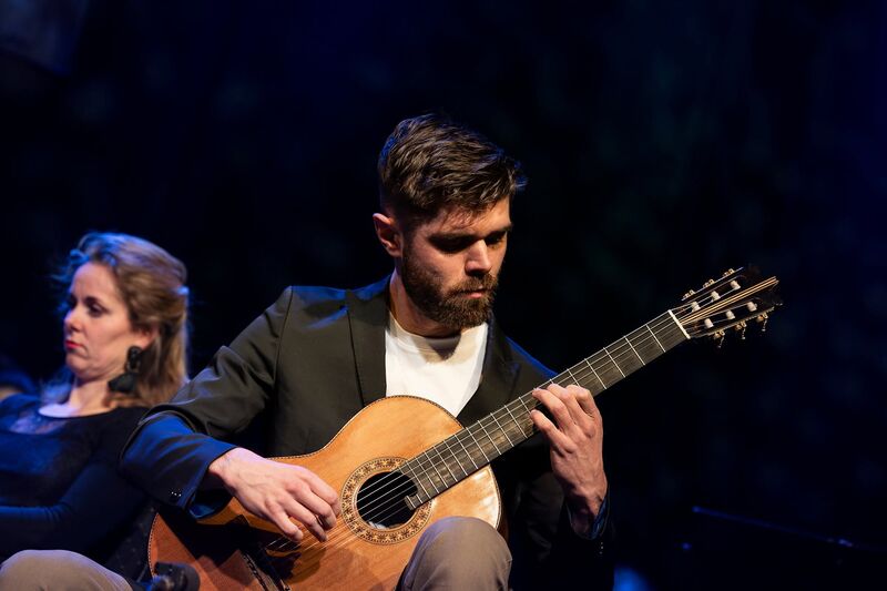 Man met een gitaar speelt op een podium, terwijl een vrouw naast hem op de achtergrond zit.