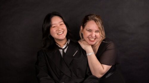 Twee vrouwen lachen samen op een donkere achtergrond, één in een zwart outfit en de ander met een lichte blouse.