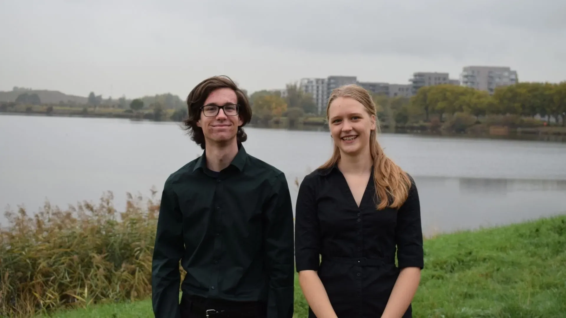 Twee mensen in zwarte kleding staan naast een meer, met gebouwen op de achtergrond en een grijzige lucht.