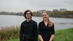 Twee mensen in zwarte kleding staan naast een meer, met gebouwen op de achtergrond en een grijzige lucht.