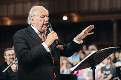 Een oudere man in een zwart vest houdt een microfoon vast en dirigeert een koor in een zaal.