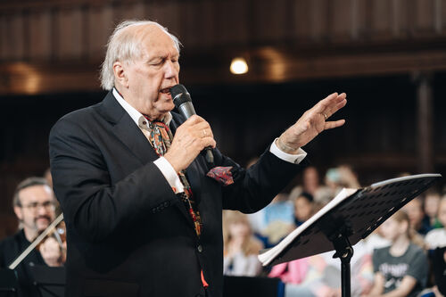 Een oudere man in een zwart vest houdt een microfoon vast en dirigeert een koor in een zaal.