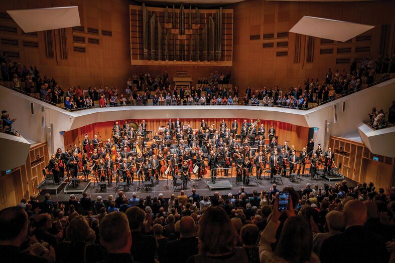 Een volle concertzaal met een orkest dat het publiek groet na een optreden.
