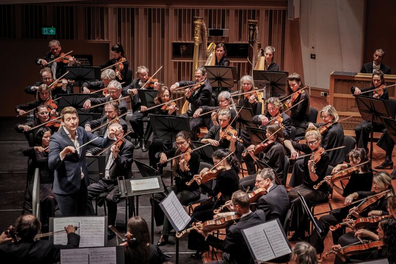Een orkest met een dirigent en musici, die samen een muziekstuk uitvoeren op het podium.
