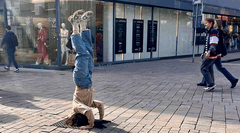 Een jongere doet een handstand op straat, terwijl omstanders voorbijlopen en winkels op de achtergrond zichtbaar zijn.