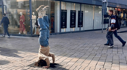 Een jongere doet een handstand op straat, terwijl omstanders voorbijlopen en winkels op de achtergrond zichtbaar zijn.