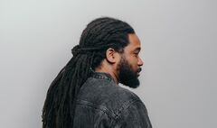 Persoon (Glen Faria) met lange dreadlocks kijkt zijwaarts terwijl hij een donkere denim jas draagt.