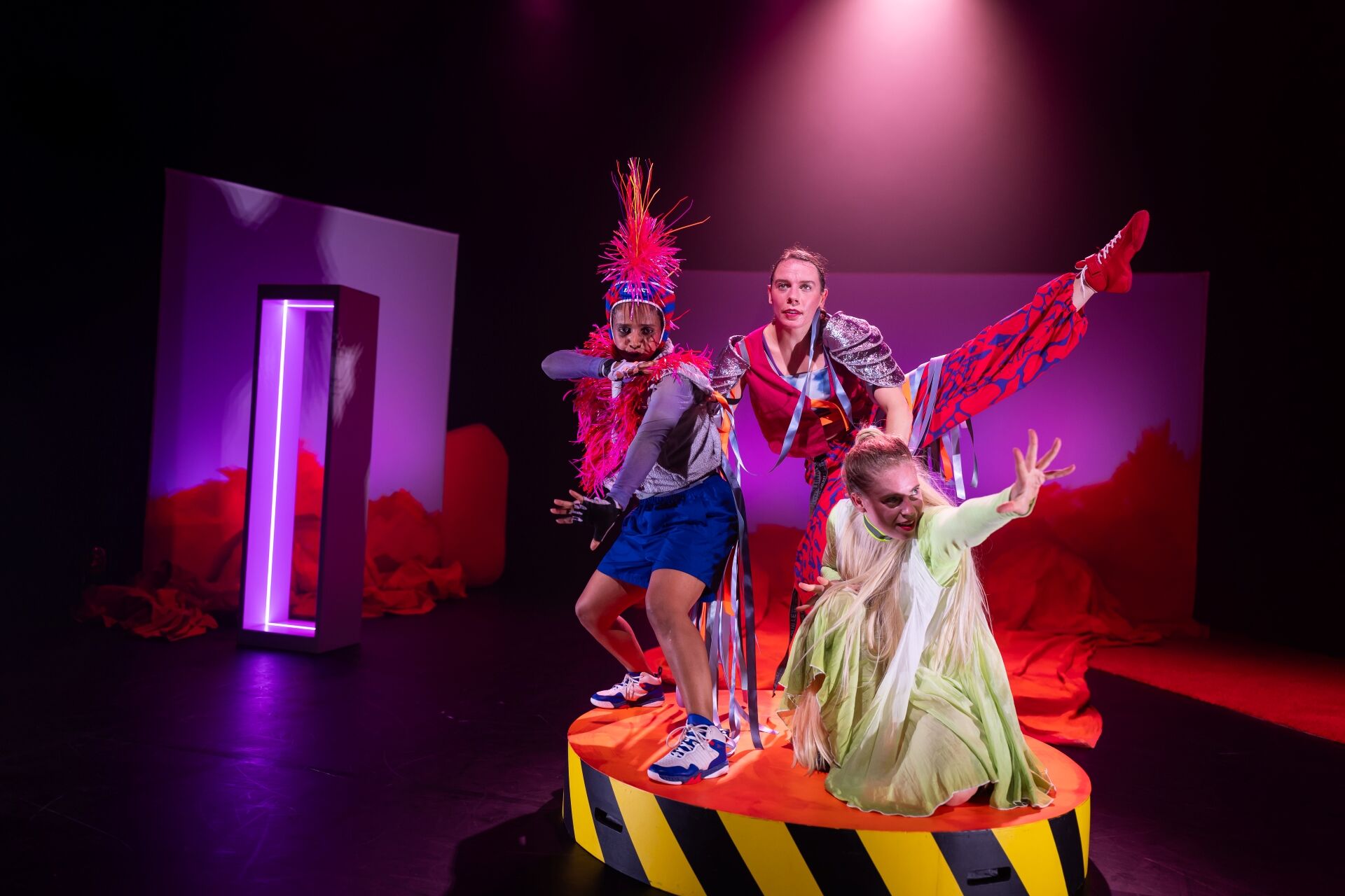 Drie performers in felgekleurde kostuums poseren op een rond platform onder theatraal licht.