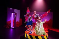 Drie performers in felgekleurde kostuums poseren op een rond platform onder theatraal licht.