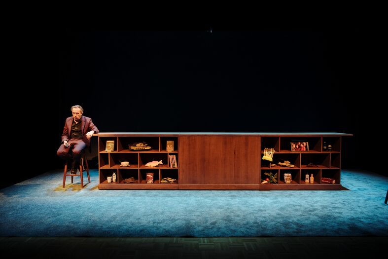 Marcel van Roosmalen zit op een barkruk naast een houten bar met planken vol voorwerpen, op een donkere theaterzaal.