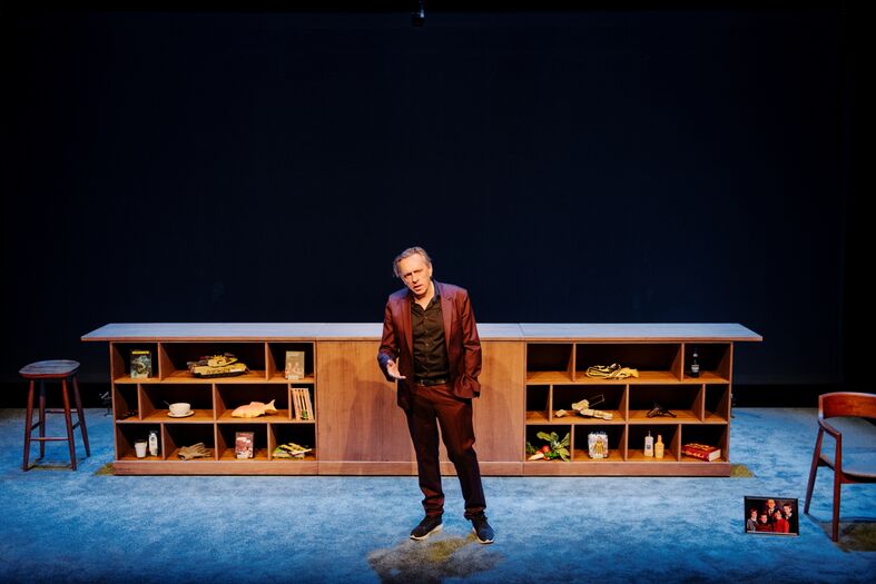 Marcel van Roosmalen in een dieprode kostuum staat op het podium voor een houten bar met verschillende accessoires.