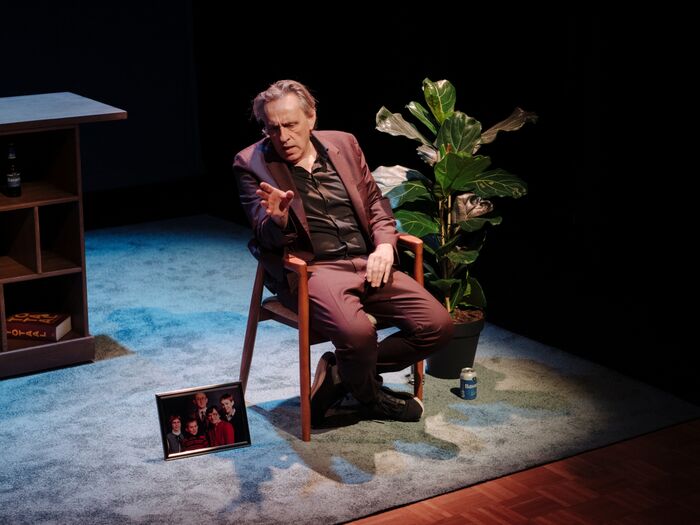 Marcel van Roosmalen in een paars pak zit op een stoel, wijst naar een foto op de grond, met een plant op de achtergrond.